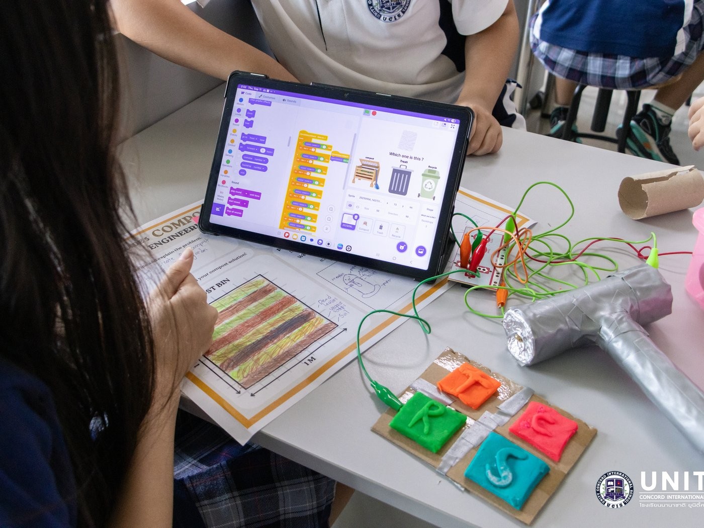 Student testing a Makey Makey and Scratch compost-sorting game they coded, next to their compost bin design worksheet
