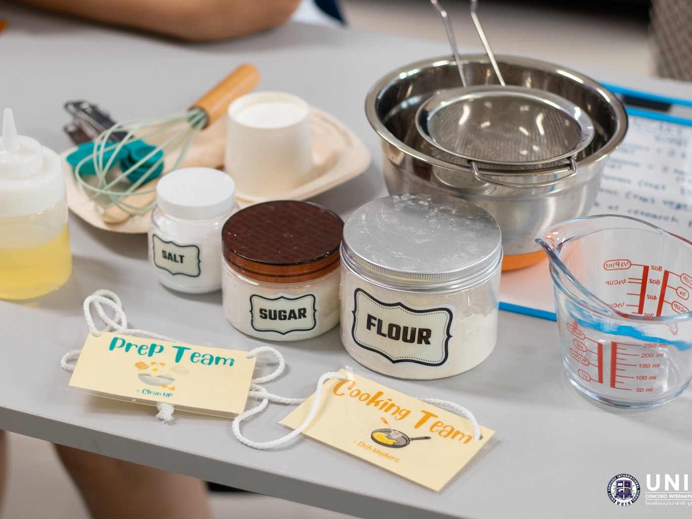 Labeled ingredient jars, a whisk, and measuring tools set out with Prep Team and Cooking Team job tags