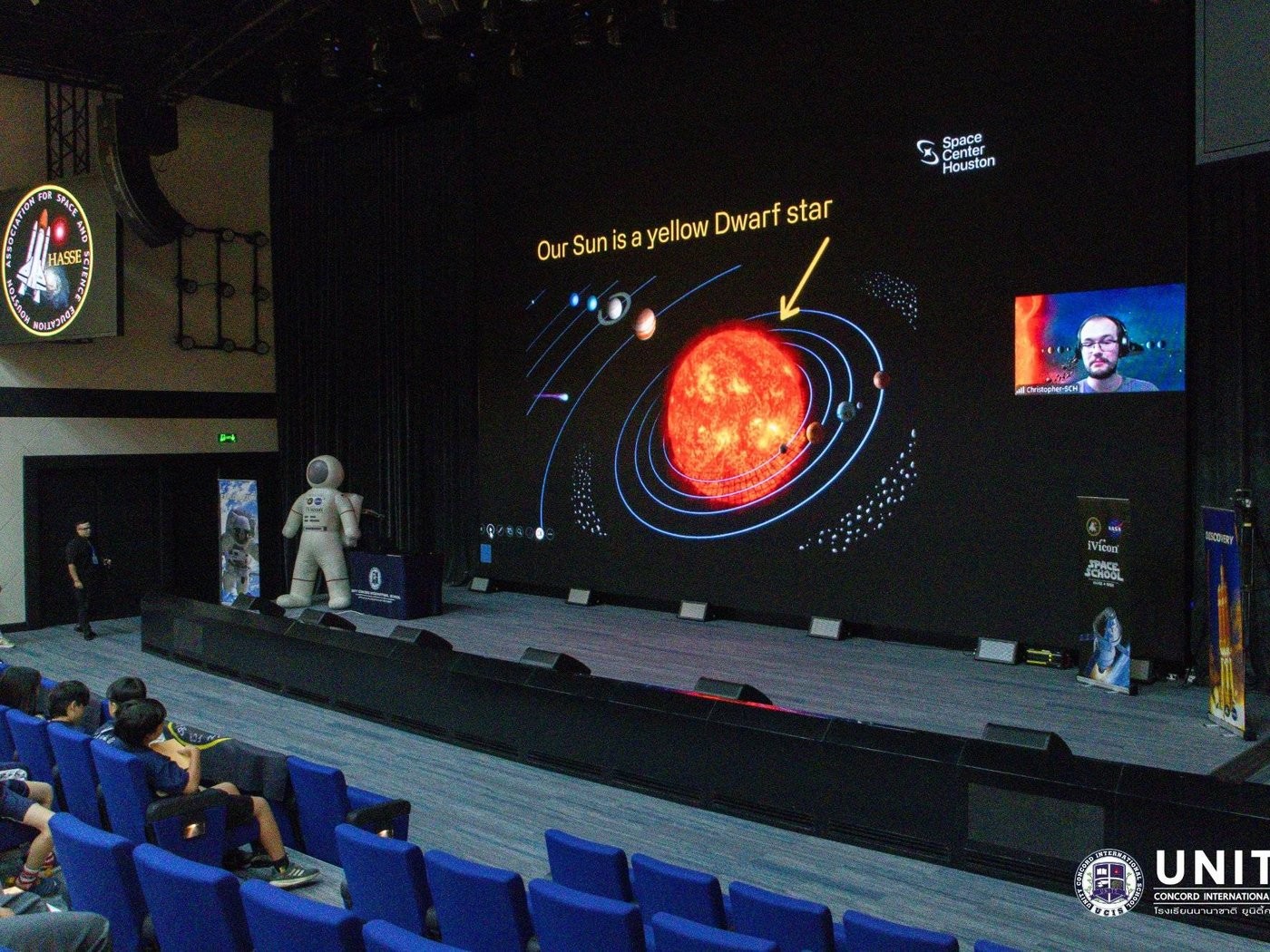 Wide view of the school auditorium during a live HASSE Space School session, with a solar system diagram on the big screen and an inflatable astronaut on stage