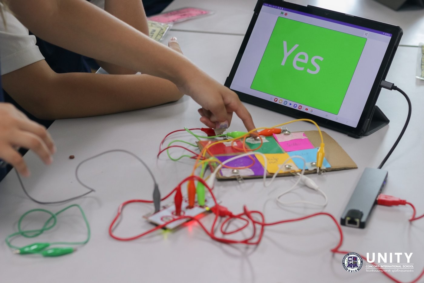 Student using a Makey Makey circuit board wired to a cardboard AAC device showing 'Yes' on a tablet
